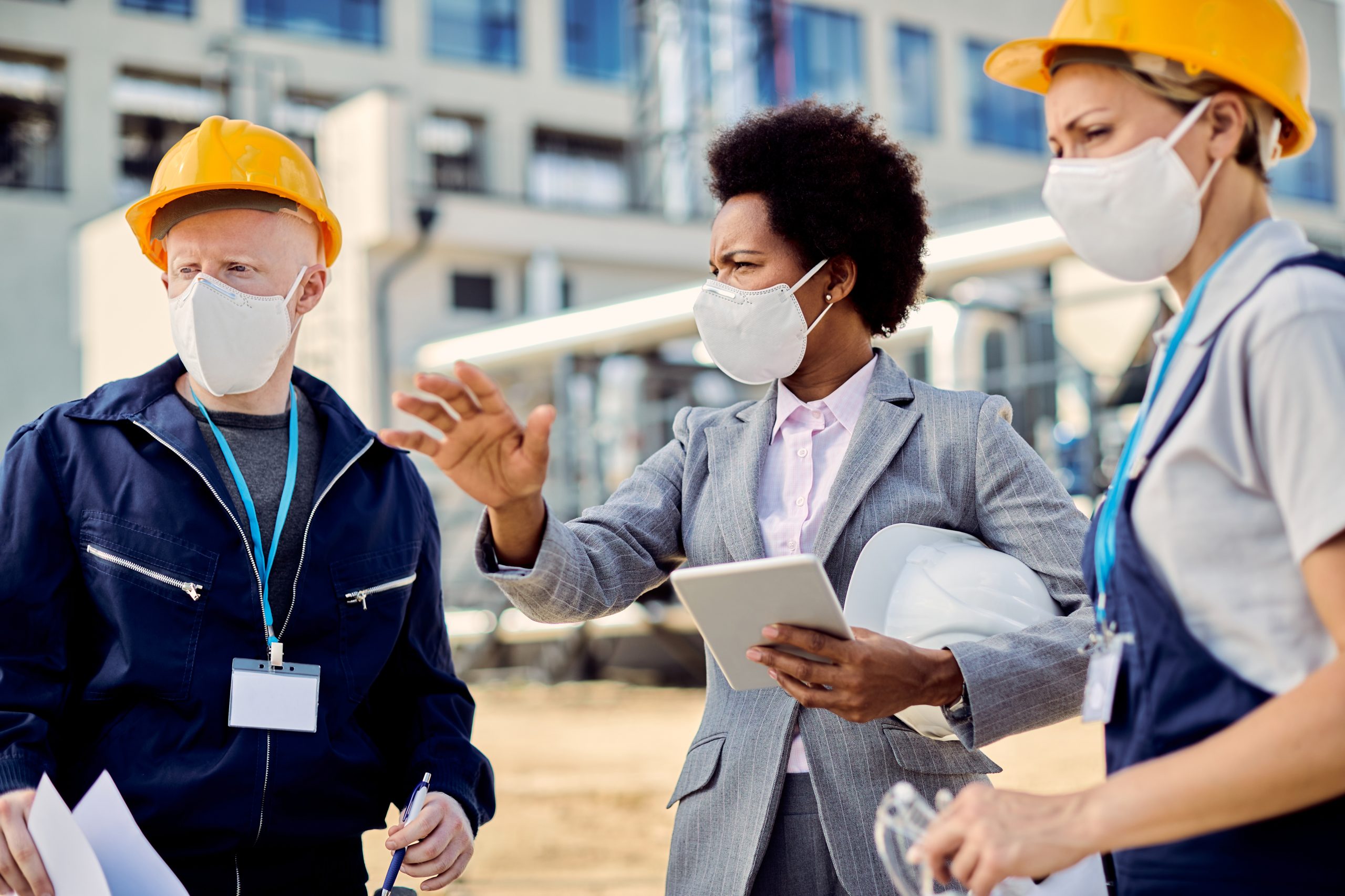 African American building contractor talking to construction engineers while inspecting project development at construction site during coronavirus epidemic.