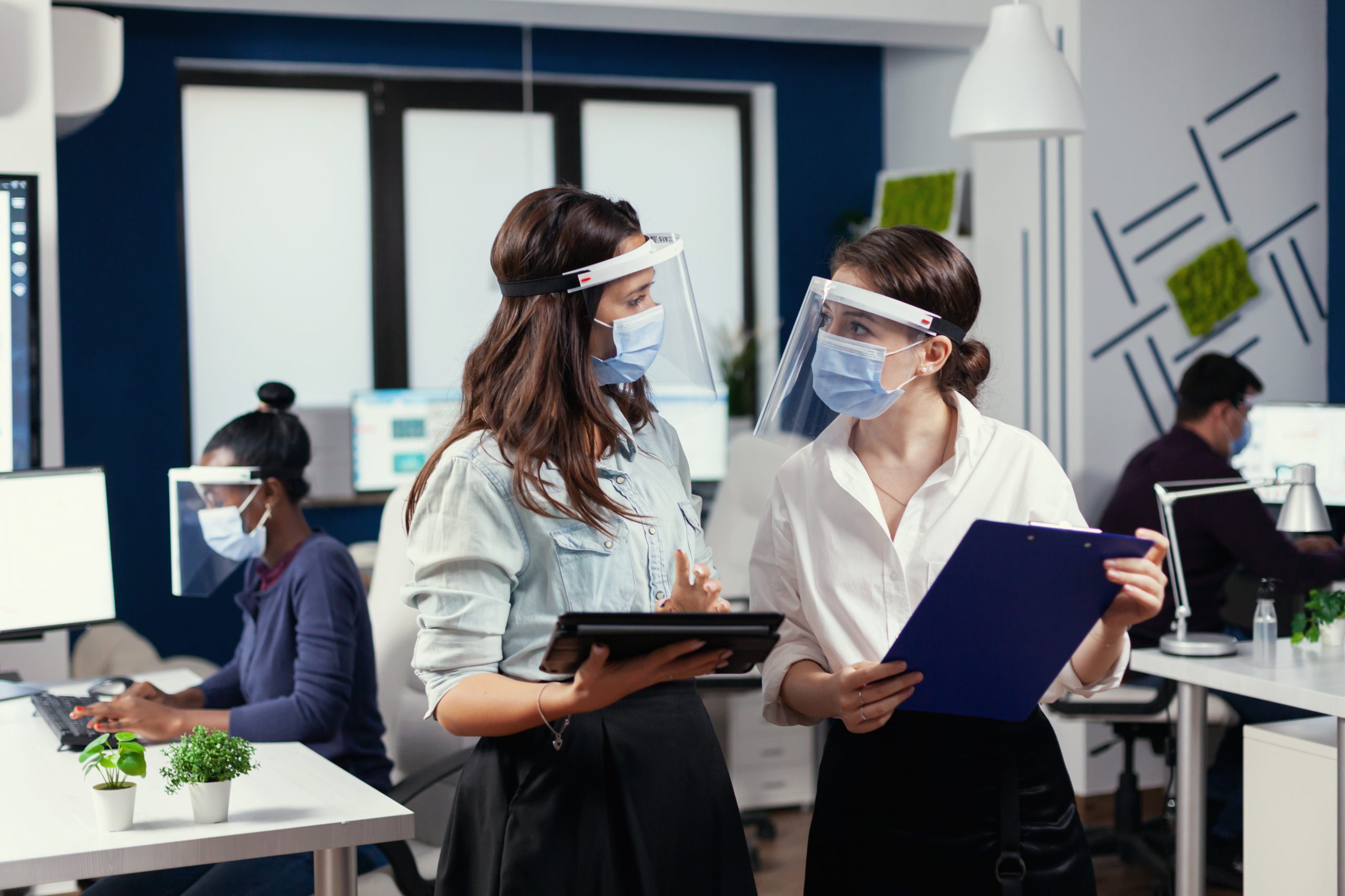 Businesswomen with protection masks against coronavirus standing in workplace talking about company financial datas holding digital tablet. Multiethnic business team working respecting social distance during global pandemic with coronavirus.