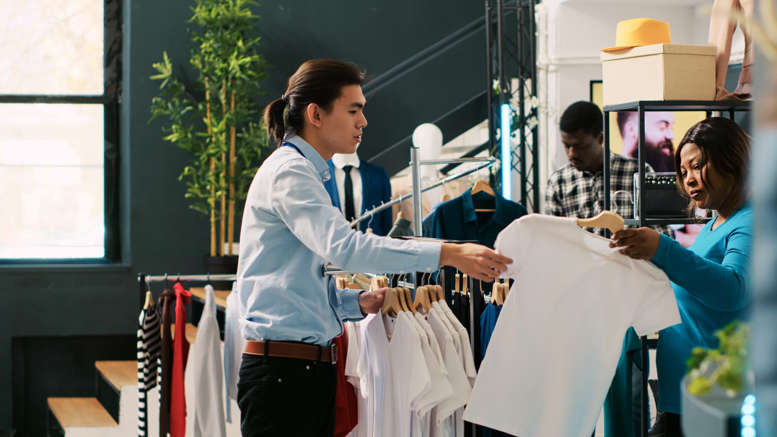 Stylish worker helping shopaholic woman with casual wear, african american woman shopping for basic clothes. Asian manager arranging fashionable merchandise in clothing store. Fashion concept